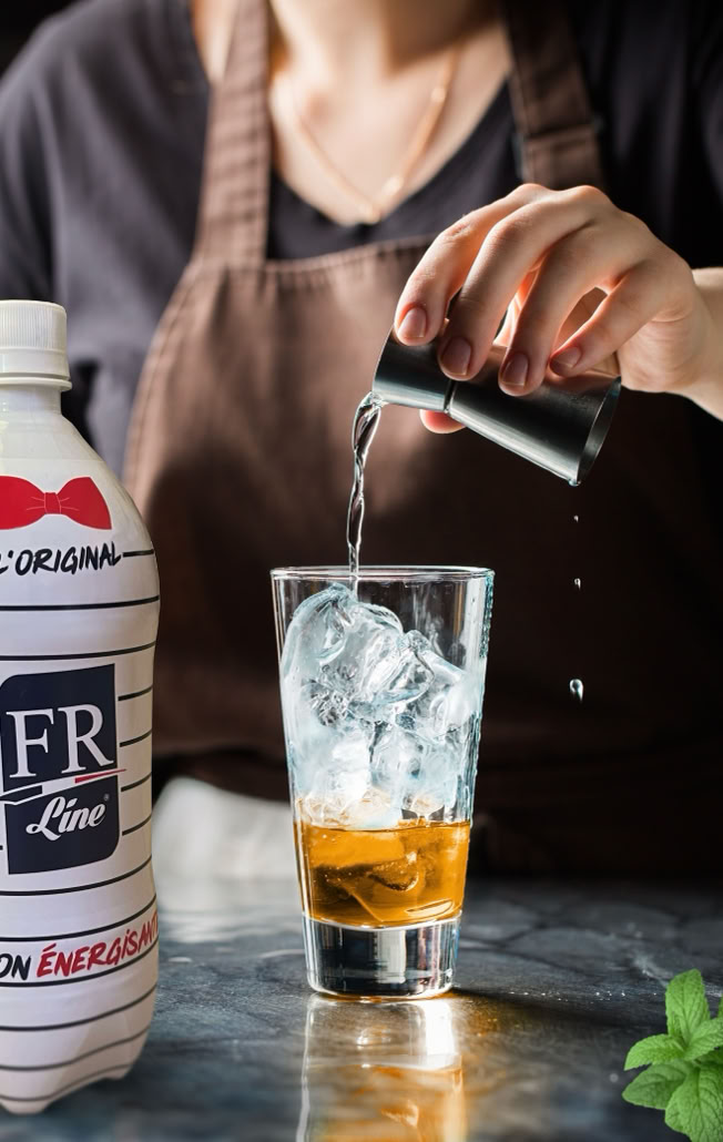 Une personne verse un liquide dans un verre rempli de glaçons, avec une bouteille blanche à gauche et des herbes vertes au premier plan.